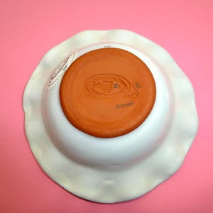 K&ouml;nnte beinhalten: Eine wei&szlig;e Keramikschale mit gewelltem Rand, von unten betrachtet. Das Innere ist in einem warmen Terrakottafarbton gehalten, mit einem gestempelten Design und dem Wort "Anismeer". Die Schale hat eine leicht unregelm&auml;&szlig;ige Form.