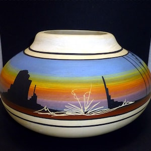 May include: A round, wide-mouthed ceramic vase featuring a hand-painted desert landscape. The design displays a gradient sky transitioning from light blue to orange and purple, with black silhouettes of rock formations and white line art of desert flora.