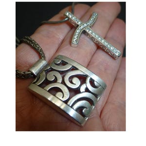 May include: A silver-toned pendant featuring an openwork scroll design on a braided chain, accompanied by a second silver-toned cross pendant inlaid with clear crystals, both suspended from a matching chain necklace.