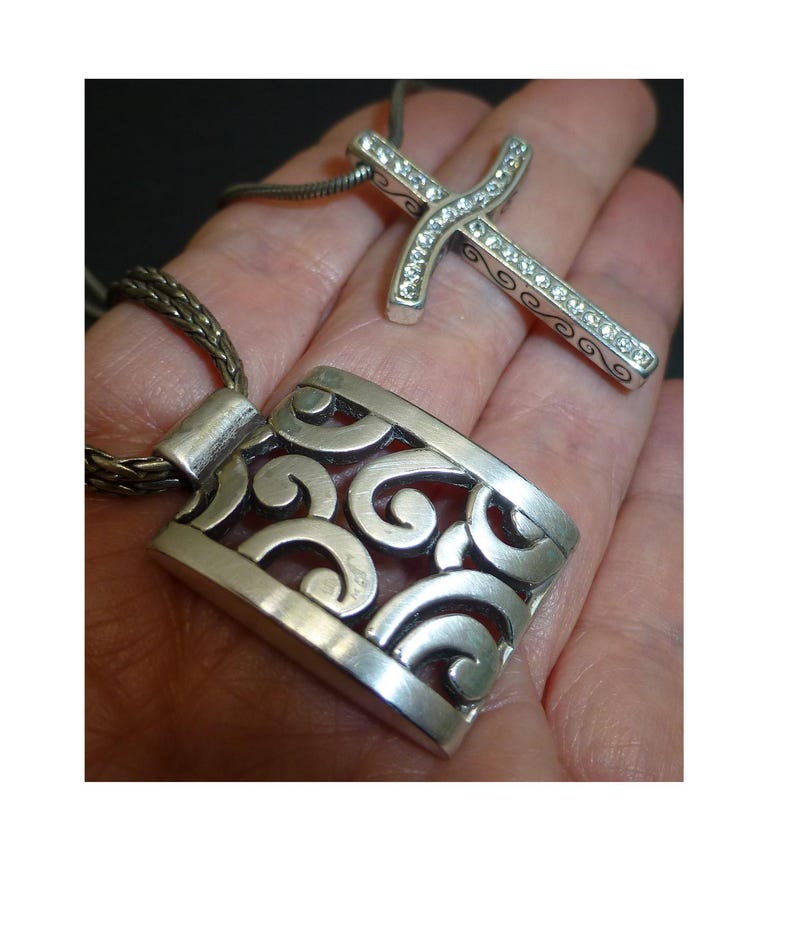 May include: A silver-toned pendant featuring an openwork scroll design on a braided chain, accompanied by a second silver-toned cross pendant inlaid with clear crystals, both suspended from a matching chain necklace.
