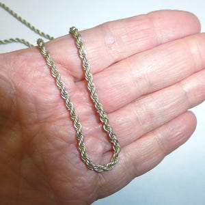 May include: A gold-colored rope chain necklace. The chain is held in a hand, and the links are tightly woven together. The necklace is a simple, elegant design.