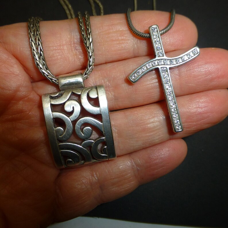 May include: Two silver-toned necklaces. One features a rectangular pendant with an openwork scroll pattern on a braided chain. The second is a cross-shaped pendant set with clear stones on a snake chain. Both pieces have a metallic finish.