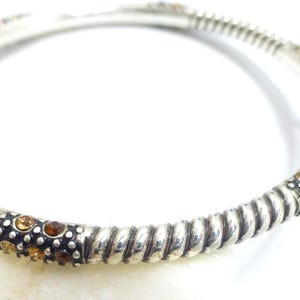 Puede incluir: Pulsera de brazalete plateada con detalles dorados. La pulsera presenta un dise&ntilde;o de cuerda trenzada y est&aacute; adornada con peque&ntilde;as piedras preciosas redondas de color &aacute;mbar. La pulsera es una joya.