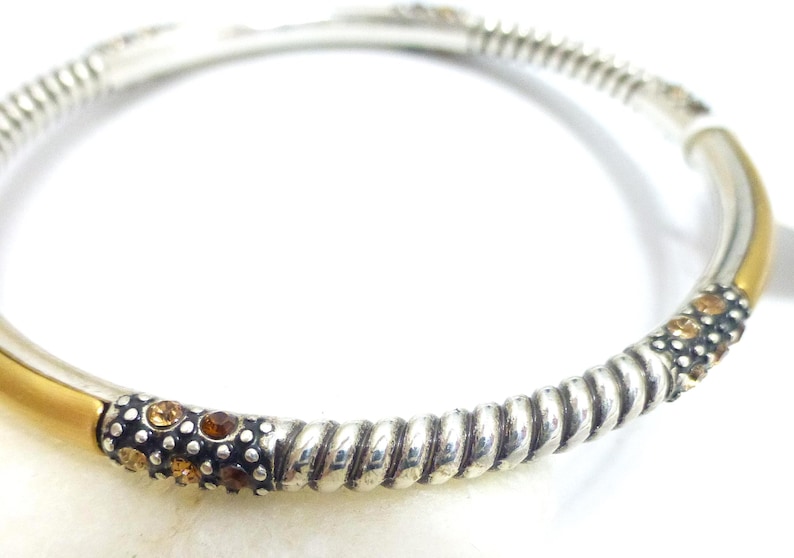 Puede incluir: Pulsera de brazalete plateada con detalles dorados. La pulsera presenta un dise&ntilde;o de cuerda trenzada y est&aacute; adornada con peque&ntilde;as piedras preciosas redondas de color &aacute;mbar. La pulsera es una joya.