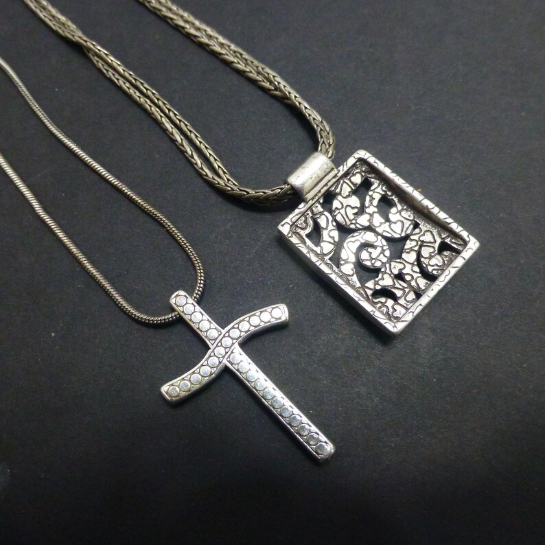 May include: Two silver-toned necklaces. One features a rectangular pendant with an intricate openwork pattern of small hearts. The other is a cross pendant decorated with a series of small circular indentations along its entire surface.