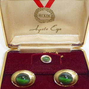 May include: Vintage gold-toned cufflinks, tie tack, and collar bar set with green agate eye stones, presented in a gold-trimmed box with a red velvet interior. The box is labeled "Hickok Agate Eye."