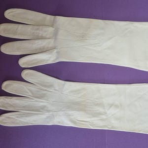 May include: A pair of long, white gloves, likely leather, are laid out on a purple surface. The gloves are smooth, with visible seams and finger detailing. The gloves appear to be in good condition.