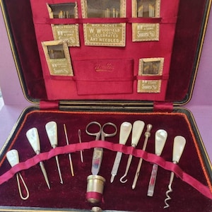 May include: A vintage sewing kit presented in a black case with a red velvet lining. The kit features tools with mother-of-pearl handles, scissors, needles, and a thimble. The case also contains packets of needles labelled "W. Woodfield & Sons, Sharps".