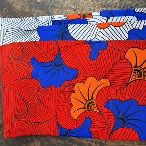 Könnte beinhalten: Ein Stapel bunter Stoffe mit einem sich wiederholenden Muster aus orangefarbenen, blauen und weißen Blumen auf rotem Hintergrund. Der Stoff ist mit "Vlisco" und "Holland" in schwarzer Schrift beschriftet.