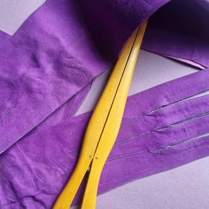May include: A pair of vintage purple leather gloves with a matching belt, and a yellow wooden glove stretcher. The gloves and belt are a vibrant purple color, with visible stitching details. The glove stretcher is a light yellow color.