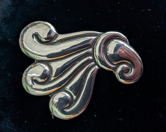May include: A silver brooch with a swirling, stylized design. The brooch features three large, curved elements that resemble stylized waves or scrolls. The metal has a polished, reflective surface.