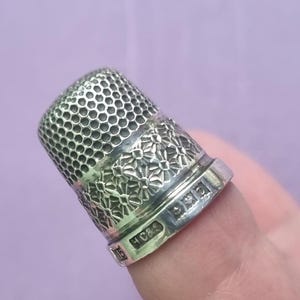 May include: A silver thimble with a floral design and a honeycomb pattern on the top. The thimble is marked with the letters "H C & S" and a diamond shape.