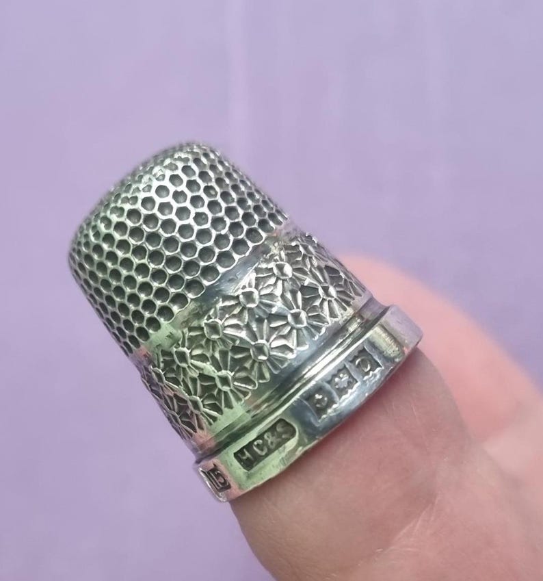 May include: A silver thimble with a floral design and a honeycomb pattern on the top. The thimble is marked with the letters "H C & S" and a diamond shape.