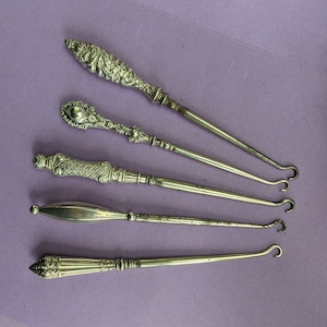 May include: Five silver shoe horn tools with ornate handles. The tools are arranged in a row on a purple surface.