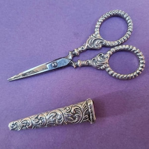 May include: An ornate pair of silver scissors with a matching sheath, set against a purple background. The scissors have intricate scrollwork and beaded finger loops. The sheath is cylindrical with similar detailing.