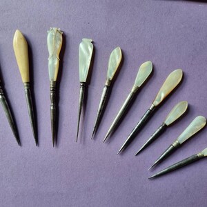 May include: A collection of nine pointed metal tools with decorative handles made of iridescent mother-of-pearl and smooth off-white material. Each tool features a slender, tapered metal shaft and a distinctively shaped handle, arranged in a fan-like formation.