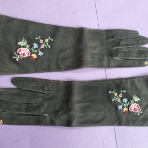 May include: A pair of long, dark green gloves with floral embroidery. The gloves feature a detailed design of pink, yellow, blue, and green flowers and leaves. The gloves are made of a soft material.