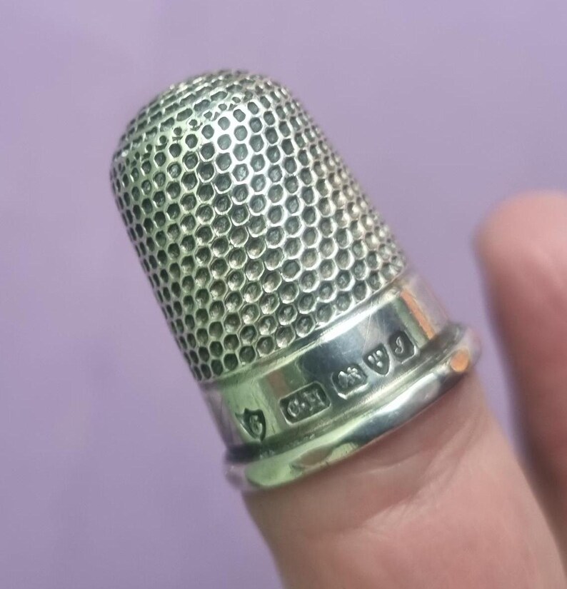 May include: A silver thimble with a textured surface and hallmarks on the base. The hallmarks include a crown, a lion, and a letter.