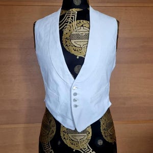 May include: A white vest with a V-neck and four buttons. The vest is displayed on a mannequin with a black and gold patterned fabric. The vest has a tailored fit and appears to be made of a lightweight material.