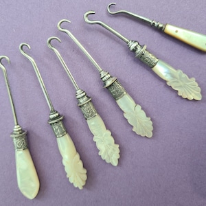 May include: Six antique silver and mother-of-pearl button hooks. The hooks are all different sizes and have intricate designs on the handles.