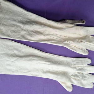 May include: A pair of off-white leather gloves with long gauntlets, laid out on a purple surface. The gloves have a textured appearance and show signs of wear on the fingertips. The gloves are designed to protect the hands and forearms.