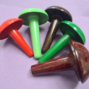 May include: Five colourful plastic bottle stoppers in the shape of mushrooms. The stoppers are red, green, brown, and black.