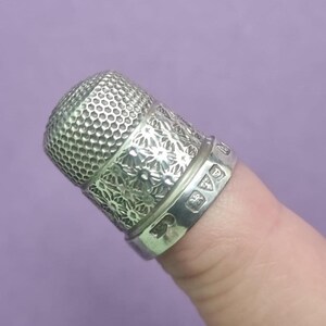 May include: A silver thimble with a floral design and a textured top. The thimble has hallmarks on the band.
