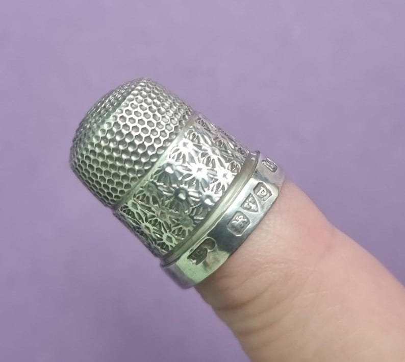 May include: A silver thimble with a floral design and a textured top. The thimble has hallmarks on the band.