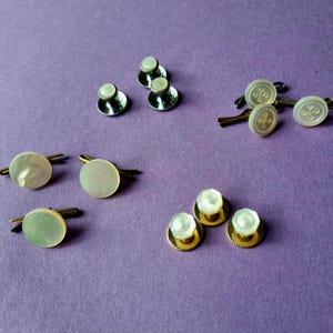 May include: A collection of cufflinks and shirt studs displayed on a purple background. The cufflinks feature round, iridescent faces with gold or silver settings. Some have a button design. The shirt studs have a similar design.