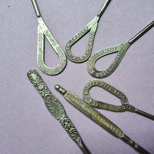 Puede incluir: Seis herramientas antiguas de metal para calzarse con diferentes lemas publicitarios. Los calzadores están hechos de metal y tienen un diseño decorativo. Los lemas incluyen "Footwear for Ladies", "With Thanks Your Chance", "Public Benefit Boot Company", "Rubber Heel Pads The Wood-MIlne", "Adamson & Barlow Bolton", y "Randalls 1008".