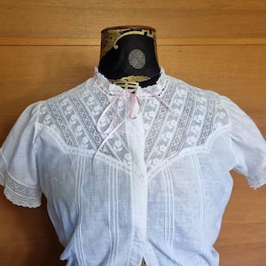 May include: White short-sleeve blouse with intricate lace detailing on the collar and yoke. The blouse features a high neckline with a delicate pink ribbon tie and vertical pleats down the front. The sleeves are trimmed with lace.