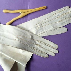 May include: A pair of off-white leather gloves with stitched details and a wooden glove stretcher on a purple surface. The gloves are long, extending past the wrist, and appear to be vintage.