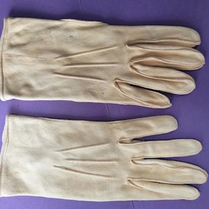 May include: A pair of light tan leather gloves. The gloves are laid flat against a purple background. Each glove has a seam running down the fingers and three parallel lines on the back of the hand.