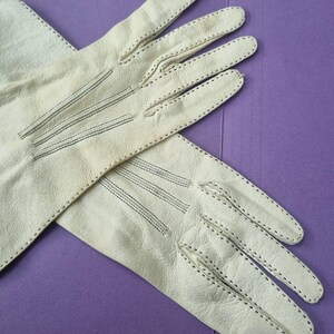 May include: A pair of off-white leather gloves with black stitching. The gloves are laid out on a purple surface. The gloves have a classic design with detailed stitching along the seams and fingers.