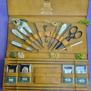 May include: Open, tan leather sewing kit with various tools, including scissors, needles, and a corkscrew. The kit contains packets of needles labeled "Sharps". The top of the case has a metal clasp and a decorative emblem.