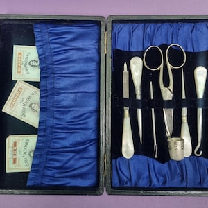 May include: A black wooden box with a blue velvet lining. The box contains a set of silver sewing tools, including a pair of scissors, a needle threader, a thimble, and a bodkin. The box also contains three cards with the text "The Sharps", "The Darners", and "The Rugs" printed on them.