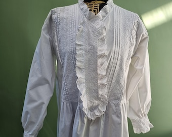 May include: White vintage dress with long sleeves. The dress features a high collar, vertical pleats, and a ruffled lace trim down the front. The bodice has detailed embroidery. The sleeves have a gathered cuff. The dress is made of a soft, flowing fabric.