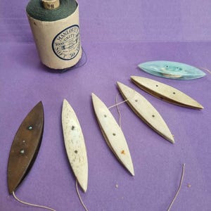 Puede incluir: Un surtido de herramientas de costura antiguas sobre una superficie morada. Incluye un carrete de hilo verde con el texto "MANLOVES UNITY", varias lanzaderas de hilo de madera y una bobina de plástico azul. Las herramientas están dispuestas para su exhibición.