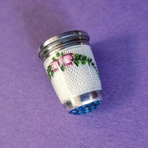 May include: A vintage thimble with a white textured body, silver trim, and a blue jeweled base. The thimble is decorated with pink floral accents and green leaves. A sewing accessory.