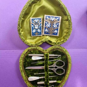 May include: A heart-shaped sewing kit case lined with green velvet, containing two blue and white floral rectangular card cases with the text 3/7, and a set of metal sewing tools including scissors, needles, and mother-of-pearl handled items.