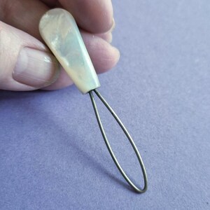 May include: A button hook tool featuring a smooth, pearlescent, off-white handle with a tapered shape and a thin, flexible metal wire loop extending from the base, designed for pulling buttons through fabric openings on garments.