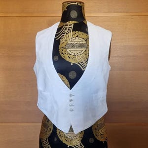 May include: A white vest with a scoop neck and three buttons. The vest is displayed on a mannequin with a black and gold patterned fabric. The mannequin is set against a wooden background.