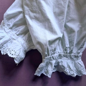 May include: A pair of white cotton bloomers featuring gathered fabric at the cuffs and a decorative eyelet lace trim along the bottom edges. The garment has a soft, opaque texture with visible pleating and intricate floral embroidery details.