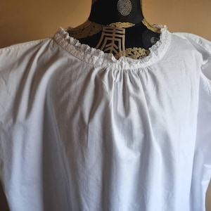 May include: A white sleeveless top featuring a gathered round neckline with a delicate lace trim. The arm openings are also finished with matching white lace edging. The garment is displayed on a black mannequin with gold patterns.