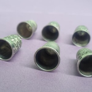 May include: Six silver thimbles with a textured pattern. The thimbles are arranged in a semi-circle on a purple surface.