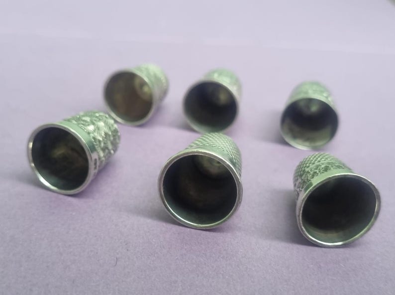 May include: Six silver thimbles with a textured pattern. The thimbles are arranged in a semi-circle on a purple surface.