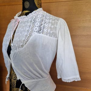 May include: White vintage blouse with lace detailing. The blouse features a high collar, short sleeves, and a delicate lace yoke. A pink ribbon adds a touch of color. The blouse is made of a lightweight fabric, perfect for layering.