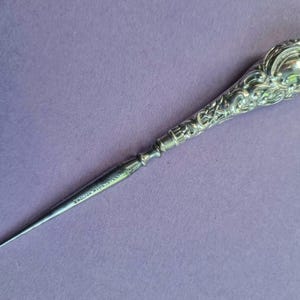 Könnte beinhalten: Verziertes silbernes Brieföffner mit detailliertem floralem Design am Griff. Die spitze Klinge trägt die Inschrift "ENGLISH MAKE". Das Objekt liegt auf einem violetten Hintergrund.