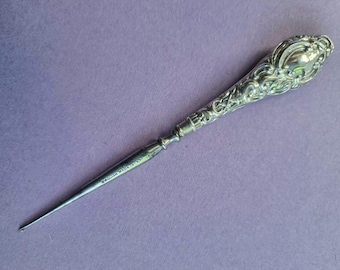 May include: Ornate silver letter opener with a detailed floral design on the handle. The pointed blade is slender, and the handle is engraved with the words "ENGLISH MAKE". The opener is displayed on a purple surface.