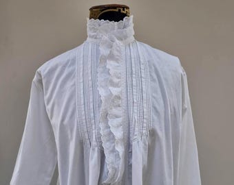 May include: A white vintage shirt with a high, ruffled collar and a pleated front. The shirt features long sleeves and delicate lace trim along the collar and down the front. The shirt is displayed on a mannequin.